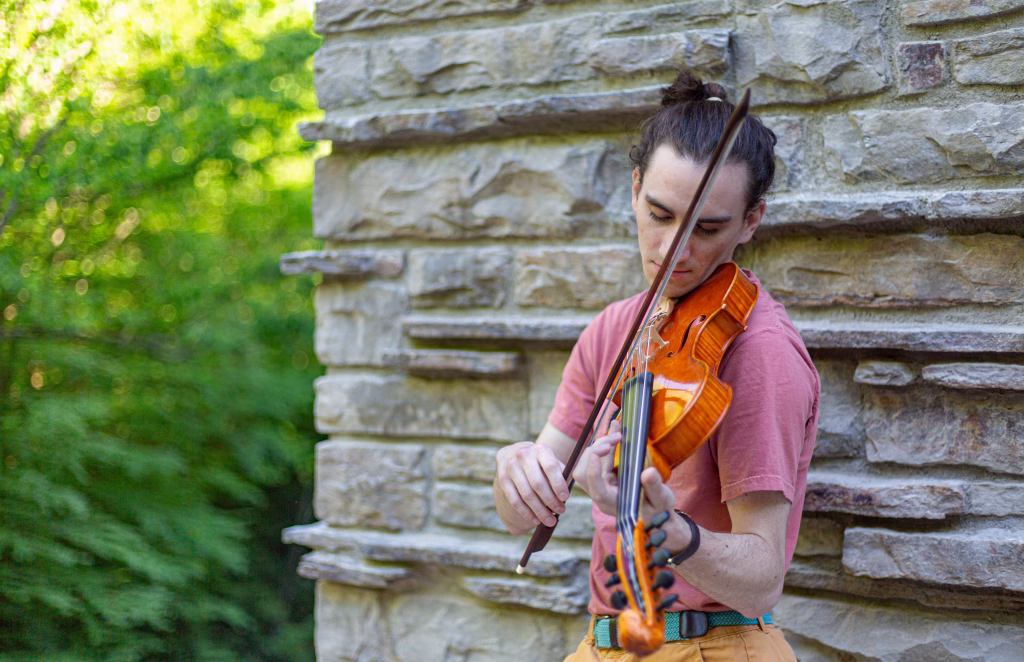 A violinist in a salmon shirt plays outdoors against a stacked stone wall backed by summer greenery.